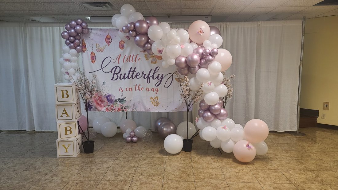 Baby shower backdrop reading A Little Butterfly is on the way, framed by a white, blush, and mauve balloon garland, blossom trees, and BABY blocks