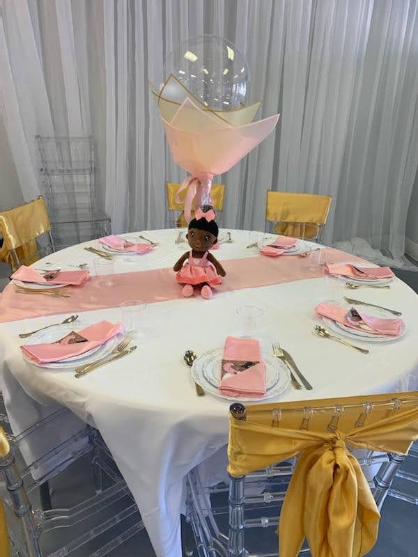 Round guest table in white, blush, and gold with a balloon bouquet centerpiece and a doll at the center